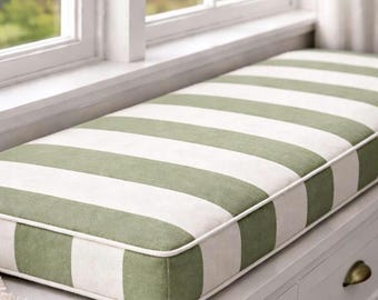 May include: A rectangular window seat cushion with wide, alternating stripes of olive green and white. The cushion has a white piped edge and is placed on a white window seat. The window is visible in the background.