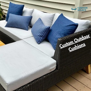 Puede incluir: Sofá seccional de exterior de mimbre marrón oscuro con cojines grises y cojines decorativos azules. El texto "Custom Outdoor Cushions" es visible en el lateral. El sofá está sobre una terraza de madera.