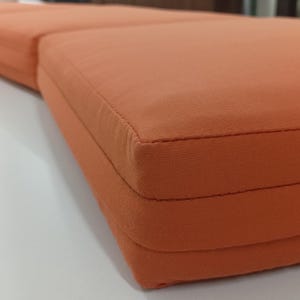 May include: Close-up of a section of an orange outdoor cushion. The cushion is made of a tightly woven fabric and has a stitched edge. The cushion is rectangular and appears to be part of a larger set.