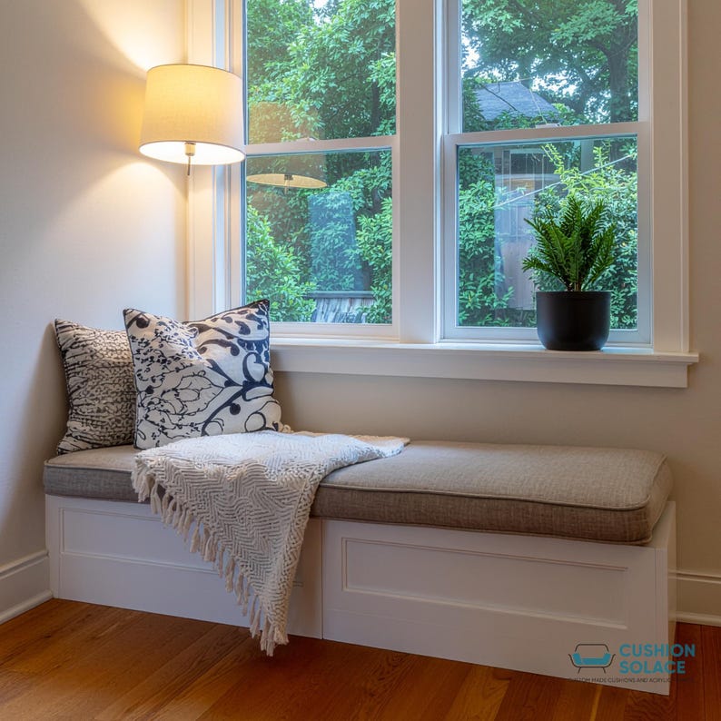 May include: A built-in window seat with a gray cushion and two decorative pillows. A white fringed throw blanket is draped over the seat. A small potted plant sits on the windowsill. A lamp is mounted on the wall.