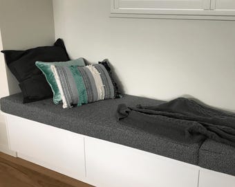 May include: A built-in bench with a gray upholstered cushion and white storage cabinets. Decorative pillows in shades of gray, teal, and white are arranged against the wall. A gray knitted throw blanket is draped over the bench. The bench is near a window with white shutters.