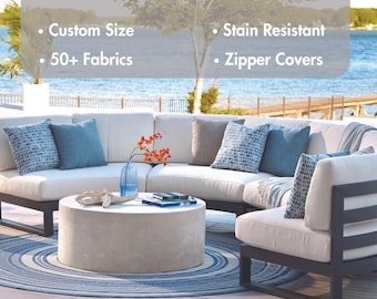 Könnte beinhalten: Ein weißes Ecksofa mit blau-weißen gemusterten Kissen, einem weißen Hocker und einem blau-weißen Teppich. Das Sofa steht auf einer Terrasse mit Blick auf ein Gewässer. Der Text "Outdoor Cushions" befindet sich oben im Bild. Der Text "Custom Size", "50+ Fabrics", "Stain Resistant" und "Zipper Covers" ist unter dem Titel aufgelistet.