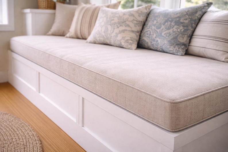 May include: A built-in window seat with a beige cushion and several decorative pillows. The pillows have various patterns and colors, including beige, blue, and white. The base of the seat is white, and the floor is hardwood.