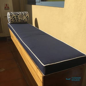 Könnte beinhalten: Ein marineblaues Outdoor-Bankkissen mit weißer Paspelierung und einem passenden Nackenkissen. Das Nackenkissen hat ein schwarz-weißes Blättermuster. Die Bank ist aus Holz und steht im Freien.
