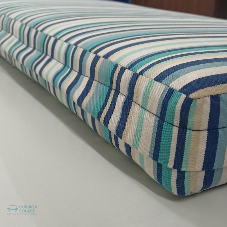 May include: Close-up of a rectangular cushion with a striped pattern in shades of blue, teal, and beige. The cushion has a thick, padded appearance, with visible seams and a soft, textured fabric. The design is reminiscent of nautical themes.