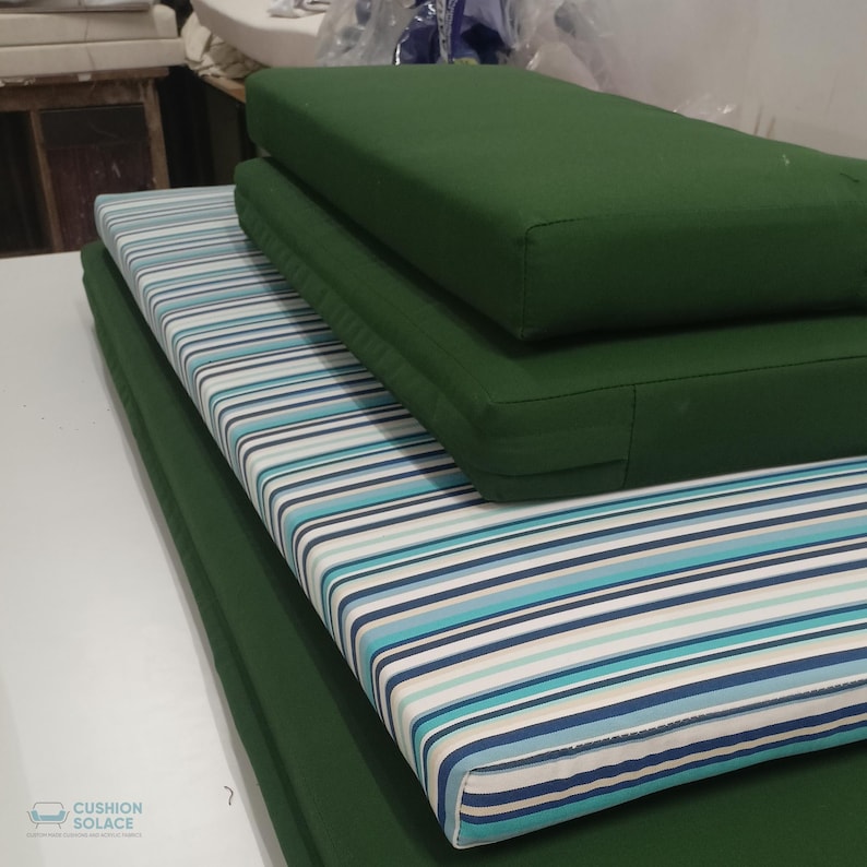 May include: A stack of outdoor cushions in various designs. The bottom cushion is solid green, followed by a striped cushion with blue, teal, and white stripes. Two solid green cushions are stacked on top. The cushions are rectangular and appear to be made of durable fabric.
