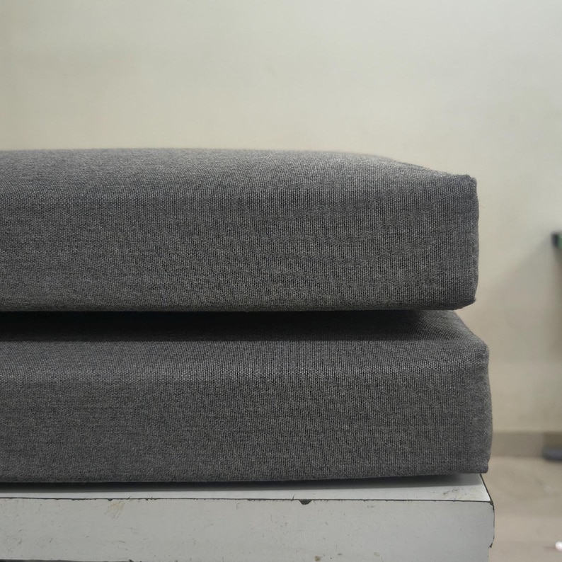 May include: Two stacked, rectangular cushions with a dark gray, textured fabric. The cushions are placed on a white surface, with a neutral background.