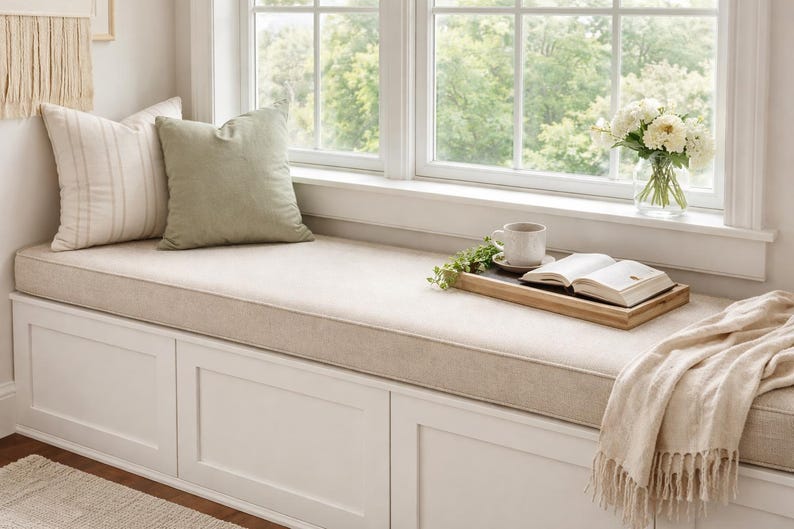 May include: A cozy window seat with a beige cushion, two pillows, and a wooden tray holding a cup, book, and greenery. A vase of white flowers sits on the windowsill. A beige throw blanket with fringe is draped on the seat.