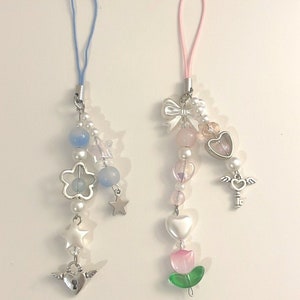 May include: Two phone charms with silver accents and beads. The charm on the left has a blue and white color scheme with a flower, stars, and a heart with wings. The charm on the right has a pink and white color scheme with a bow, a heart, a key with wings, and a pink flower.