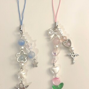 May include: Two phone charms with colorful beads, charms, and a string for hanging. One charm has a blue string and features a flower, stars, and a heart with wings. The other charm has a pink string and features a bow, a heart, a key with wings, and a pink flower.