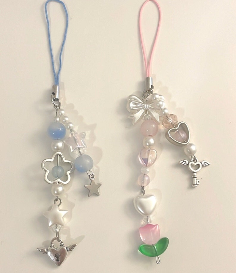 May include: Two phone charms with colorful beads, charms, and a string for hanging. One charm has a blue string and features a flower, stars, and a heart with wings. The other charm has a pink string and features a bow, a heart, a key with wings, and a pink flower.