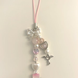 May include: A pink and white phone charm with a bow, beads, a heart, a key with wings, and a green flower.