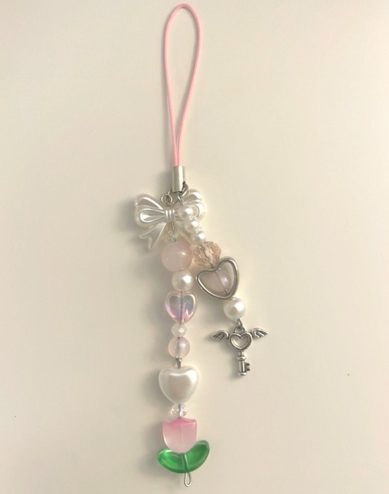 May include: A pink and white phone charm with a bow, beads, a heart, a key with wings, and a green flower.