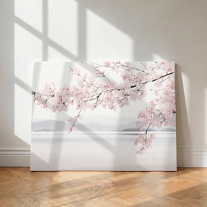 May include: A canvas print depicting a tranquil scene with cherry blossom branches. The artwork presents pale pink flowers against a soft white backdrop, with a suggestion of mountains. The print is displayed on a wooden floor.