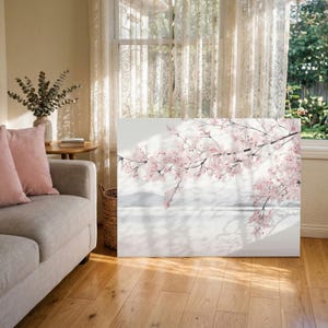 Puede incluir: Impresión en lienzo de una rama de flores de cerezo rosas sobre un fondo blanco. La luz del sol proyecta sombras suaves sobre la obra de arte. La impresión se exhibe en un salón luminoso con suelo de madera y un sofá.