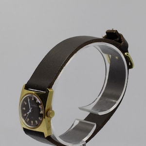 Puede incluir: Un reloj de pulsera dorado con una esfera negra y una correa de cuero marrón. El reloj está en un expositor transparente.