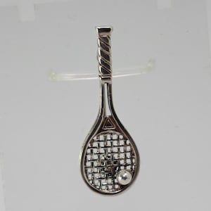 Pode incluir: Pingente de raquete de tênis prateada com uma bola. A raquete tem um cabo detalhado e um design de rede na cabeça. A bola é uma pequena esfera prateada. O pingente é provavelmente feito de prata esterlina.