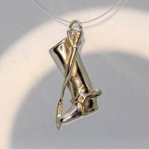 Może przedstawiać: Srebrny wisiorkowy zawieszek przedstawiający kozak jeździecki ze ostrogą. Kozak jest ozdobiony klamrą i szwami.