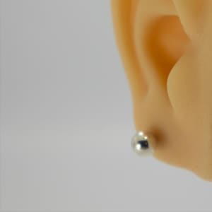 Op de afbeelding: Een kleine, glanzende, zilverkleurige baloorbel is zichtbaar in de oorlel van een persoon. De oorbel heeft een eenvoudig, rond knopontwerp en reflecteert licht. De achtergrond is effen van kleur.