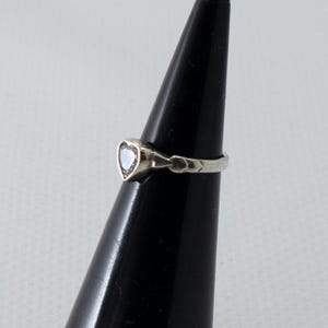 Puede incluir: Un anillo de plata con una piedra iridiscente en forma de corazón. El anillo se exhibe sobre un soporte negro en forma de cono, sobre un fondo neutro. La piedra en forma de corazón es el punto focal del anillo.