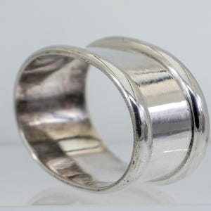 Könnte beinhalten: Ein silberfarbener Ring mit breitem Band und vertieftem Mittelteil. Der Ring hat eine glatte, polierte Oberfläche und ein schlichtes, elegantes Design. Der Ring ist wahrscheinlich aus Sterlingsilber oder einem ähnlichen Metall gefertigt.