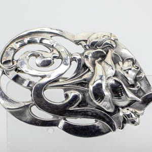 以下が含まれることがあります： 銀色の、アールヌーボー様式のブローチ。流れるようなライン、花のモチーフ、そして詳細な顔が特徴です。金属は光を反射し、奥行きを出す、磨かれた仕上げです。装飾的でエレガントな作品です。