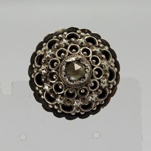 以下が含まれることがあります： 中央に灰色の宝石をあしらった、詳細なアンティークシルバーボタン。ボタンは、宝石の周りに円と星型の複雑な透かし彫りデザインが施されています。ボタンの縁は暗いです。