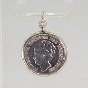 May include: A silver pendant featuring a coin with a profile of a woman and the text "WILHELMINA KONINGIN DER NEDERLANDEN". The coin is set in a circular frame with a small loop for a chain.