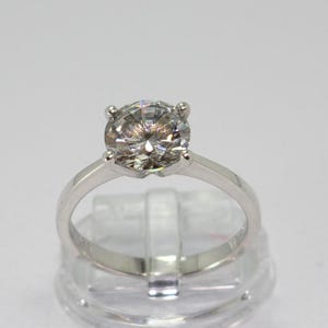 Op de afbeelding: Een zilveren verlovingsring met een grote, ronde, heldere edelsteen. De edelsteen is gezet in een vier-klauwzetting en de band is glad en gepolijst. De ring wordt gepresenteerd op een transparante plastic standaard.