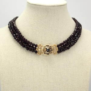 Peut inclure: Collier à plusieurs rangs avec des perles rouge foncé. Le collier est doté d'un fermoir doré avec une pierre bleu foncé au centre. Le collier est présenté sur un mannequin.
