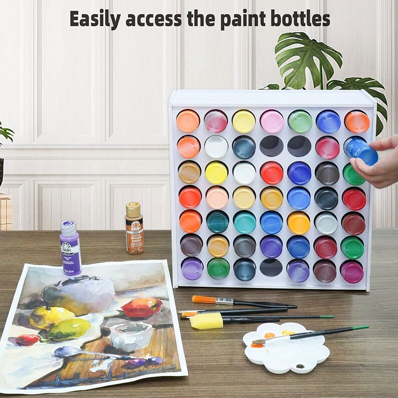 May include: White paint bottle storage rack with 48 slots. The rack is filled with various colors of paint bottles. A person is holding a blue paint bottle. The rack is on a wooden table with a painting, brushes, and a paint palette.