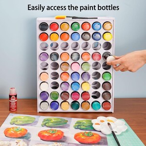 May include: White paint bottle storage rack with 72 individual slots. The rack is filled with various colored paint bottles. A hand is reaching into the rack to grab a bottle. The text "Easily access the paint bottles" is at the top of the image.