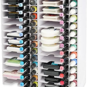 May include: A white, multi-compartment organizer filled with various art supplies. It contains ink pads, markers, and ink bottles in a range of colors. The organizer is designed to hold and display art supplies, keeping them organized and accessible.