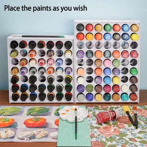 May include: Two white paint storage racks with rows of colorful paint bottles. The racks are on a table with a painting, a paint palette, and brushes.