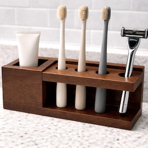 May include: A dark brown wooden bathroom organiser holding a tube of toothpaste, three toothbrushes, and a razor. The toothbrushes are beige and grey. The razor is silver and black. The organiser sits on a speckled countertop.