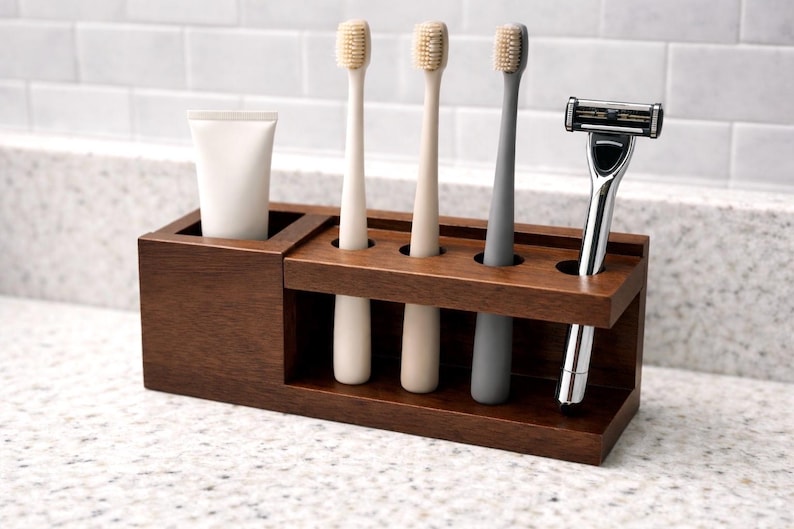 May include: A dark brown wooden bathroom organiser holding a tube of toothpaste, three toothbrushes, and a razor. The toothbrushes are beige and grey. The razor is silver and black. The organiser sits on a speckled countertop.
