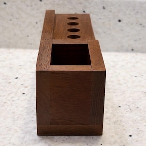 May include: A dark brown wooden desk tidy. It features a square base with a rectangular opening and a top section with four circular holes. The design is simple and functional, suitable for office or home use, with measurements in centimetres.