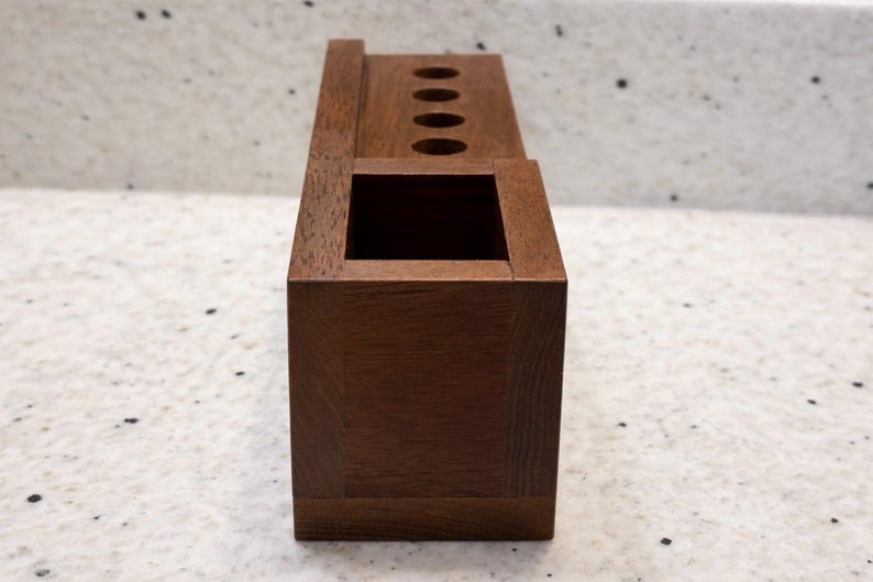 May include: A dark brown wooden desk tidy. It features a square base with a rectangular opening and a top section with four circular holes. The design is simple and functional, suitable for office or home use, with measurements in centimetres.