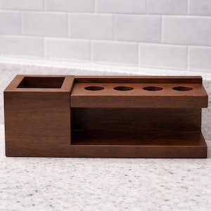 May include: A dark brown wooden desk organiser. It features a square compartment and a section with three circular holes. The organiser has a shelf underneath the holes and is made of solid wood. The unit is designed for desk organisation.