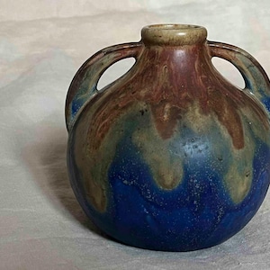 Peut inclure: Un petit vase en céramique bleu et brun avec deux anses. Le vase a un glaçage marbré avec des nuances de bleu, de brun et de beige.