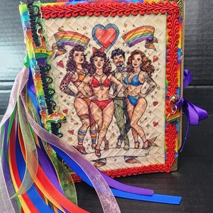 May include: A decorative journal with a colorful cover featuring a vintage-style illustration of four figures with rainbow flags and hearts. The journal is adorned with red trim, colorful ribbons, and a rainbow charm.