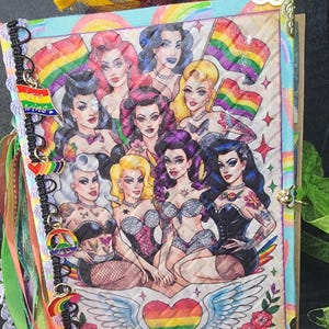 May include: A handmade journal with a colorful illustration of drag queens, rainbow flags, and a heart with wings. The journal has a decorative spine with charms and ribbons. The cover features a gold border and a clasp.