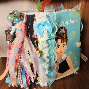 May include: A handmade journal featuring a cover with an illustration of Audrey Hepburn. The journal is adorned with lace, ribbons, and a blue bow. Decorative beads and charms are attached to the side, adding to its unique, vintage aesthetic.