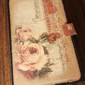 May include: A vintage journal with a black cover and a floral design. The cover has a faded, aged appearance, featuring pink roses and a decorative lace border. The journal includes a small tab closure.