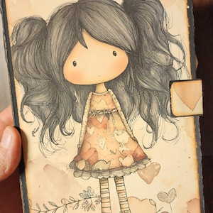 May include: A hand-drawn illustration of a doll-like figure with long, dark hair in pigtails. The doll wears a peach-coloured dress with heart patterns and striped stockings. The artwork has a vintage, storybook aesthetic, with a small envelope-shaped closure.
