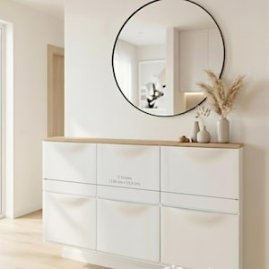 Könnte beinhalten: Moderne Eingangseinrichtung mit einem großen, runden Spiegel mit schwarzem Rahmen über einem weißen Schrank mit heller Holzplatte. Der Schrank hat sechs Türen und Schubladen. Der Text "3 Trones (154 cm x 15,9 cm)" ist sichtbar. Dekorative Vasen und Trockenblumen stehen auf dem Schrank.