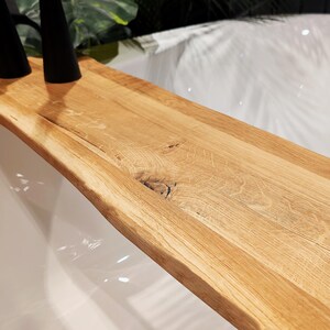May include: A natural wood bath tray with a live edge design. The tray is made of light-colored wood and has a curved shape. Two black objects are on the tray. The tray is placed over a white bathtub.
