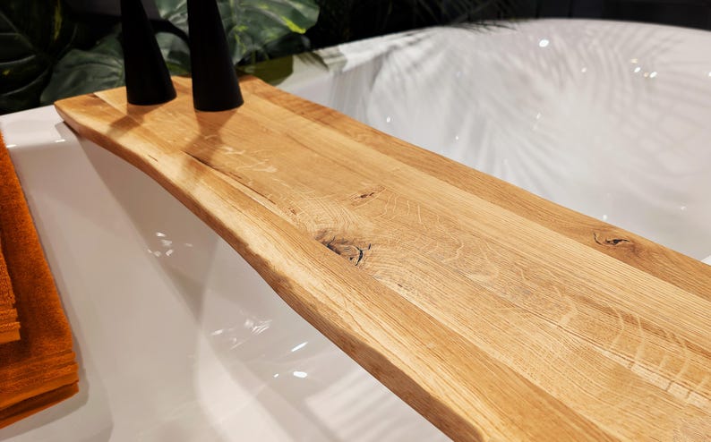 May include: A natural wood bath tray with a live edge design. The tray is made of light-colored wood and has a curved shape. Two black objects are on the tray. The tray is placed over a white bathtub.