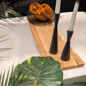 May include: A wooden tray holds a black wire basket with orange towels and two black candle holders with white candles. Green leaves and a white surface are in the foreground.