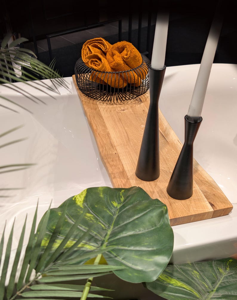 May include: A wooden tray holds a black wire basket with orange towels and two black candle holders with white candles. Green leaves and a white surface are in the foreground.